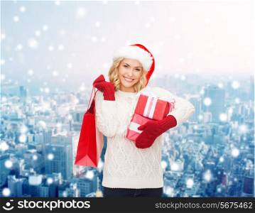 happiness, winter holidays, christmas and people concept - smiling young woman in santa helper hat with gifts and shopping bag over snowy city background