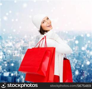 happiness, winter holidays, christmas and people concept - smiling young woman in white hat and mittens with red shopping bags over snowy city background