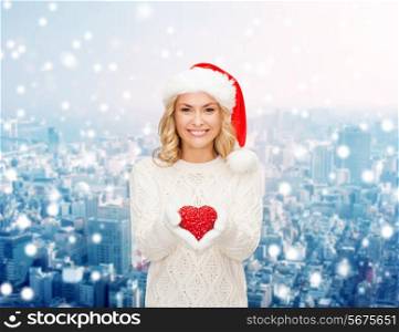 happiness, winter holidays, christmas and people concept - smiling young woman in santa helper hat with red heart decoration over snowy city background