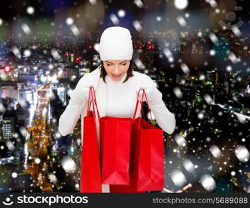 happiness, winter holidays, christmas and people concept - smiling young woman in white hat and mittens with red shopping bags over snowy city night background