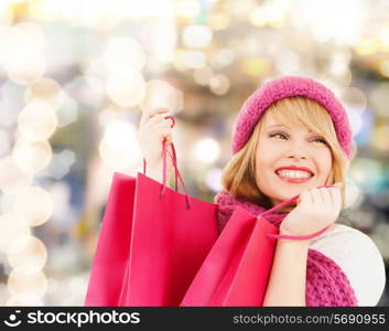 happiness, winter holidays, christmas and people concept - smiling young woman in hat and scarf with pink shopping bags over lights background