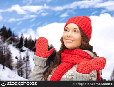 happiness, winter holidays, christmas and people concept - smiling young woman in red hat, scarf and mittens over snowy mountains background