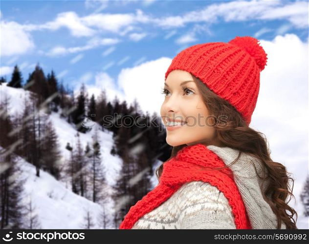 happiness, winter holidays, christmas and people concept - smiling young woman in red hat and scarf over snowy mountains background