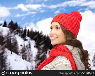 happiness, winter holidays, christmas and people concept - smiling young woman in red hat and scarf over snowy mountains background