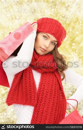 happiness, winter holidays, christmas and people concept - smiling young woman in hat and scarf with shopping bags over yellow lights background