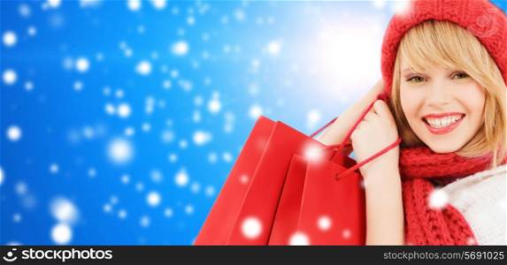 happiness, winter holidays, christmas and people concept - smiling young woman in hat and scarf with shopping bags over blue snowy background