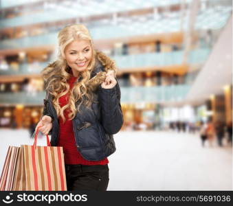 happiness, winter holidays, christmas and people concept - smiling young woman in winter clothes with bags over shopping center background