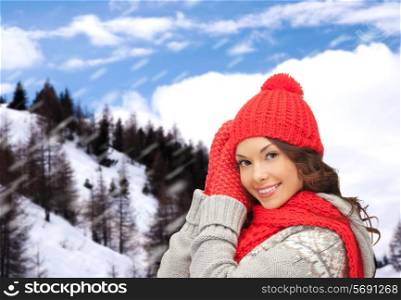 happiness, winter holidays, christmas and people concept - smiling young woman in red hat, scarf and mittens over snowy mountains background