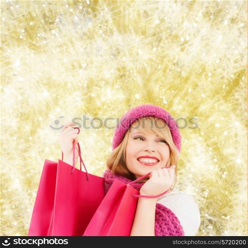 happiness, winter holidays, christmas and people concept - smiling young woman in hat and scarf with pink shopping bags over yellow lights background
