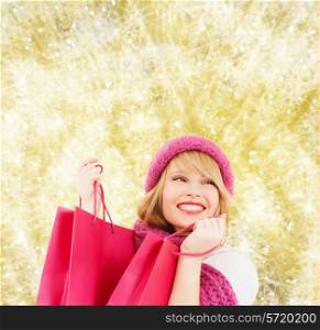 happiness, winter holidays, christmas and people concept - smiling young woman in hat and scarf with pink shopping bags over yellow lights background