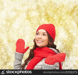 happiness, winter holidays, christmas and people concept - smiling young woman in red hat, scarf and mittens over yellow lights background