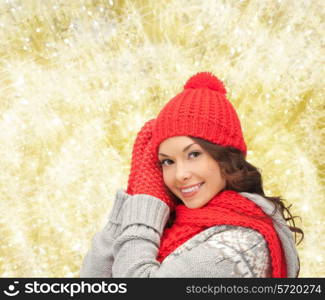 happiness, winter holidays, christmas and people concept - smiling young woman in red hat, scarf and mittens over yellow lights background