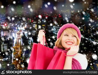 happiness, winter holidays, christmas and people concept - smiling young woman in hat and scarf with pink shopping bags ove snowy night city background