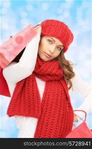 happiness, winter holidays, christmas and people concept - smiling young woman in hat and scarf with shopping bags over blue lights background
