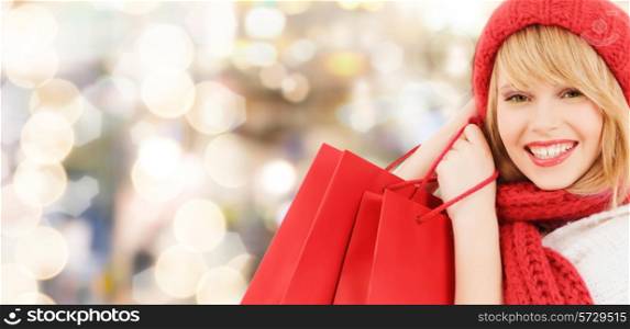 happiness, winter holidays, christmas and people concept - smiling young woman in hat and scarf with red shopping bags over lights background