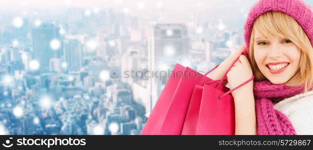 happiness, winter holidays, christmas and people concept - smiling young woman in hat and scarf with pink shopping bags ove snowy city background