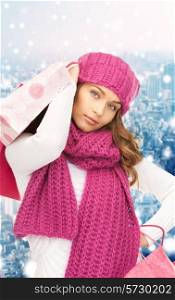 happiness, winter holidays, christmas and people concept - smiling young woman in hat and scarf with shopping bags over snowy city background
