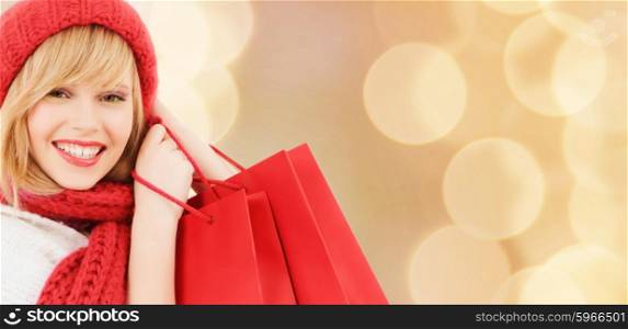 happiness, winter holidays, christmas and people concept - smiling young woman in hat and scarf with red shopping bags over beige lights background