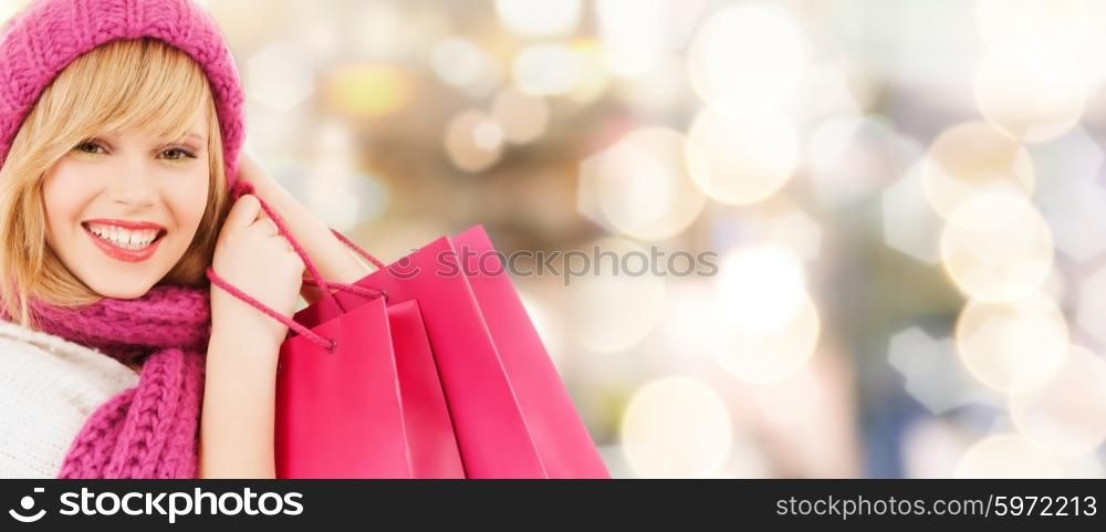 happiness, winter holidays, christmas and people concept - smiling young woman in hat and scarf with pink shopping bags over lights background