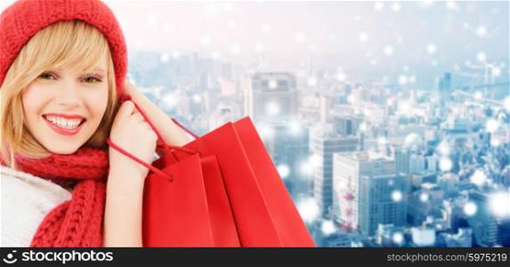 happiness, winter holidays, christmas and people concept - smiling young woman in hat and scarf with shopping bags over snowy city background