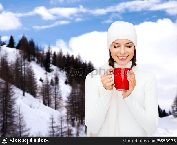 happiness, winter holidays, christmas, beverages and people concept - smiling young woman in white warm clothes with red cup over snowy mountains background