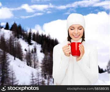 happiness, winter holidays, christmas, beverages and people concept - smiling young woman in white warm clothes with red cup over snowy mountains background