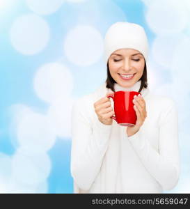 happiness, winter holidays, christmas, beverages and people concept - smiling young woman in white warm clothes with red cup over blue lights background