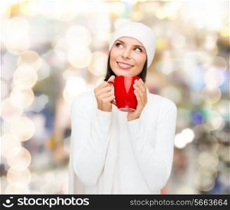 happiness, winter holidays, christmas, beverages and people concept - smiling young woman in white warm clothes with red cup over lights background