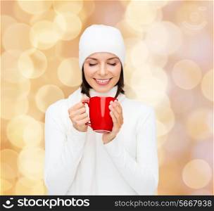 happiness, winter holidays, christmas, beverages and people concept - smiling young woman in white warm clothes with red cup over beige lights background