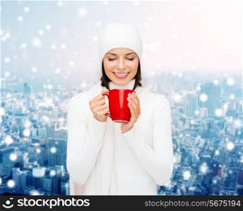 happiness, winter holidays, christmas, beverages and people concept - smiling young woman in white warm clothes with red cup over snowy city background