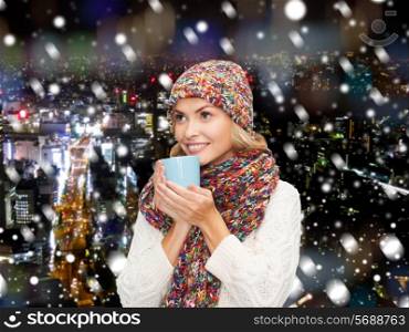 happiness, winter holidays, christmas, beverages and people concept - smiling young woman in warm clothes with cup over snowy city background