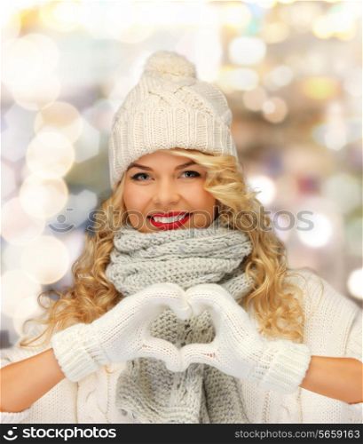 happiness, winter holidays, gesture, charity and people concept - smiling young woman in white hat and mittens showing heart-shape gesture over shiny lights background