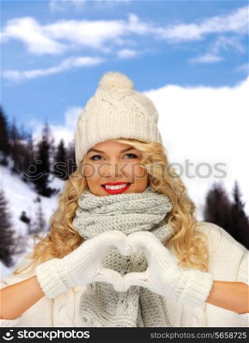 happiness, winter holidays, tourism, travel and people concept - smiling young woman in white hat and mittens showing heart-shape gesture over snowy mountains background