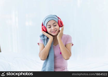 Happy a asian women disease mammary cancer patient with pink ribbon wearing headscarf in headphones is listening to music After treatment to chemotherapy sit on bed In the bedroom at the house