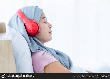 Happy a asian women disease mammary cancer patient with pink ribbon wearing headscarf in headphones is listening to music After treatment to chemotherapy sit on bed In the bedroom at the house