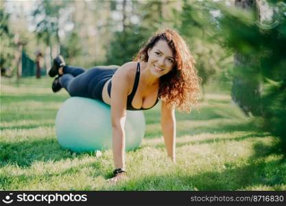 Happy active sportswoman does fitness exercises with gymnastic ball, dressed in active wear, has workout in open air, poses on green grass in forest or park, focused somewhere with pleasant smile