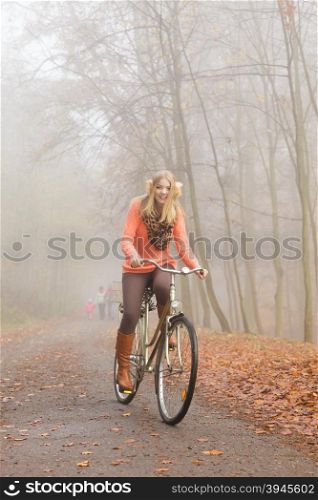 Happy active woman riding bike in autumn park.. Happy active woman riding bike bicycle in foggy fall autumn park. Glad young girl in sweater and earmuffs relaxing. Healthy lifestyle and recreation leisure activity.