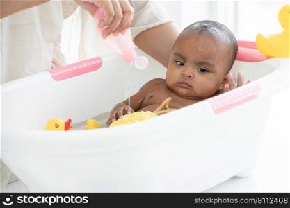 Happy adorable African newborn baby bathing in bathtub. Mother pouring shower gel on sponge to wash her little daughter in warm water. Newborn baby cleanliness care concept
