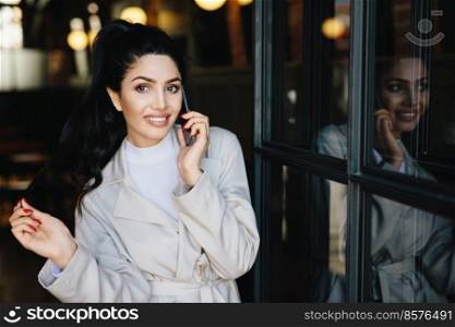 Happy adorable brunette woman with appealing appearance dressed in white clothes speaking over cell phone gossiping with her friend. Young businesswoman communicating over modern mobile phone
