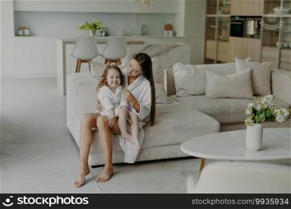 Happy adorable little hands poses on mothers legs dressed in soft white bath towel feels very glad. Mom and daughter wear domestic clothes sit on comfortable sofa in living room. Home interior