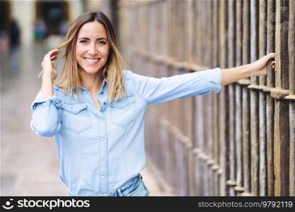 Happy adult female in stylish blue shirt looking at camera with smile and touching hair while grasping metal fence on blurred background of city street. Cheerful woman touching hair near fence
