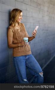 Happy adult female with coffee to go smiling and watching video on cellphone while leaning on gray wall on city street. Cheerful woman with smartphone and coffee leaning on wall
