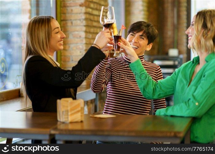Happy adult girlfriends smiling and clinking glasses of alcohol drinks while standing near table and window during meeting in bar. Excited women proposing toast near pub window