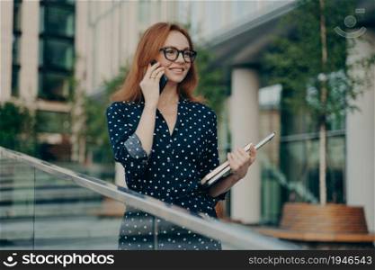 Happy adult lady with red hair makes consultancy call on smartphone spends leisure for international talking wears spectacles polka dot dress holds diary poses outdoor in urban setting focused forward. Happy adult lady with red hair makes consultancy call on smartphone