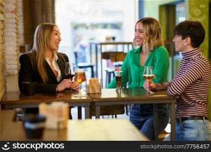 Happy adult women with alcohol drinks smiling and talking while gathering around table during friends meeting in pub. Cheerful girlfriends gathering around table in bar
