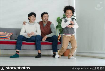 Happy African adopted kid boy wearing sweater dancing Hip Hop in front of Asian young gay parent looking with pride face at living room at home. Lgbt family supporting dream of their son