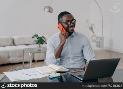 Happy african american businessman makes business call, enjoying mobile conversation, working at laptop at workplace. Smiling black male manager discussing job by phone, sitting at modern office desk.. African american businessman makes phone business call enjoying mobile conversation works at laptop