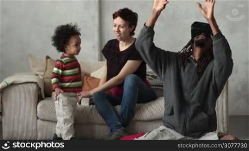 Happy african american father playing with virtual reality goggles headset glasses at home. Mixed race curly toddler son imitating his moves with hands while smiling caucasian mother sitting on sofa and laughing. People using new trends technology.