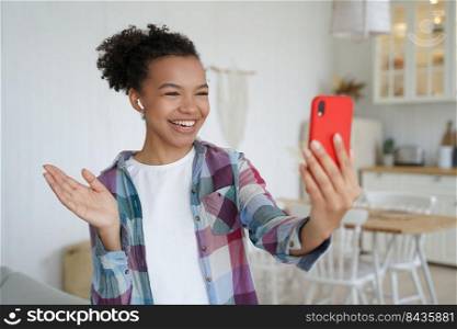 Happy african american girl in wireless earphone holds smartphone, talking on video call, waving hand hello. Friendly smiling young female blogger chatting online with friends at home.. Happy african american girl blogger hold smartphone, talking on videocall, waving hand hello at home