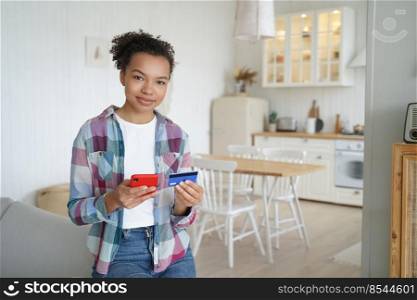 Happy african american girl is buying online with mobile phone and plastic credit card from home. Teenager spends money. Online shopping with credit card, internet banking. Business and e-commerce.. Happy african american girl is buying online with mobile phone and credit card from home.
